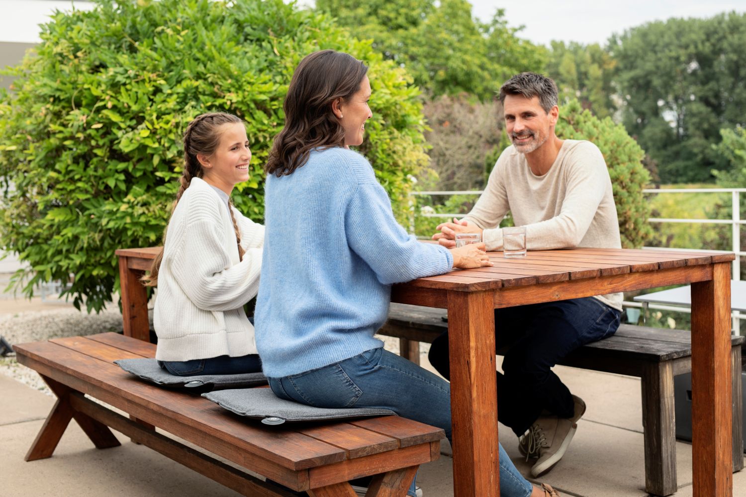 Eine Familie sitzt an einem Holztisch im Garten und nutzt beheizte Sitzkissen auf der Sitzbank.